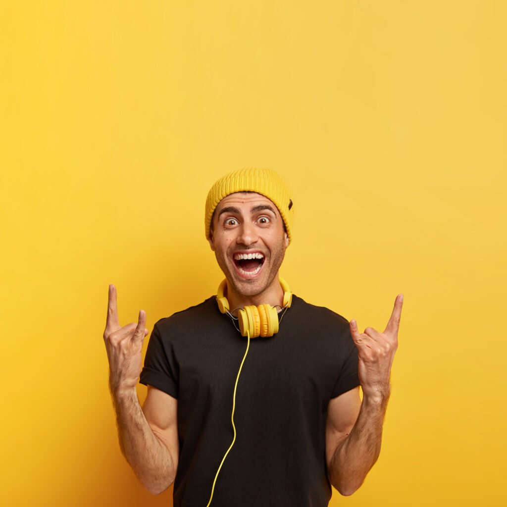 carefree joyful guy makes rock n roll gesture, brings positive vibes, listens rock music in headphones, enjoys cool sound, wears stylish yellow hat and black t shirt, stands on yellow wall, copy space