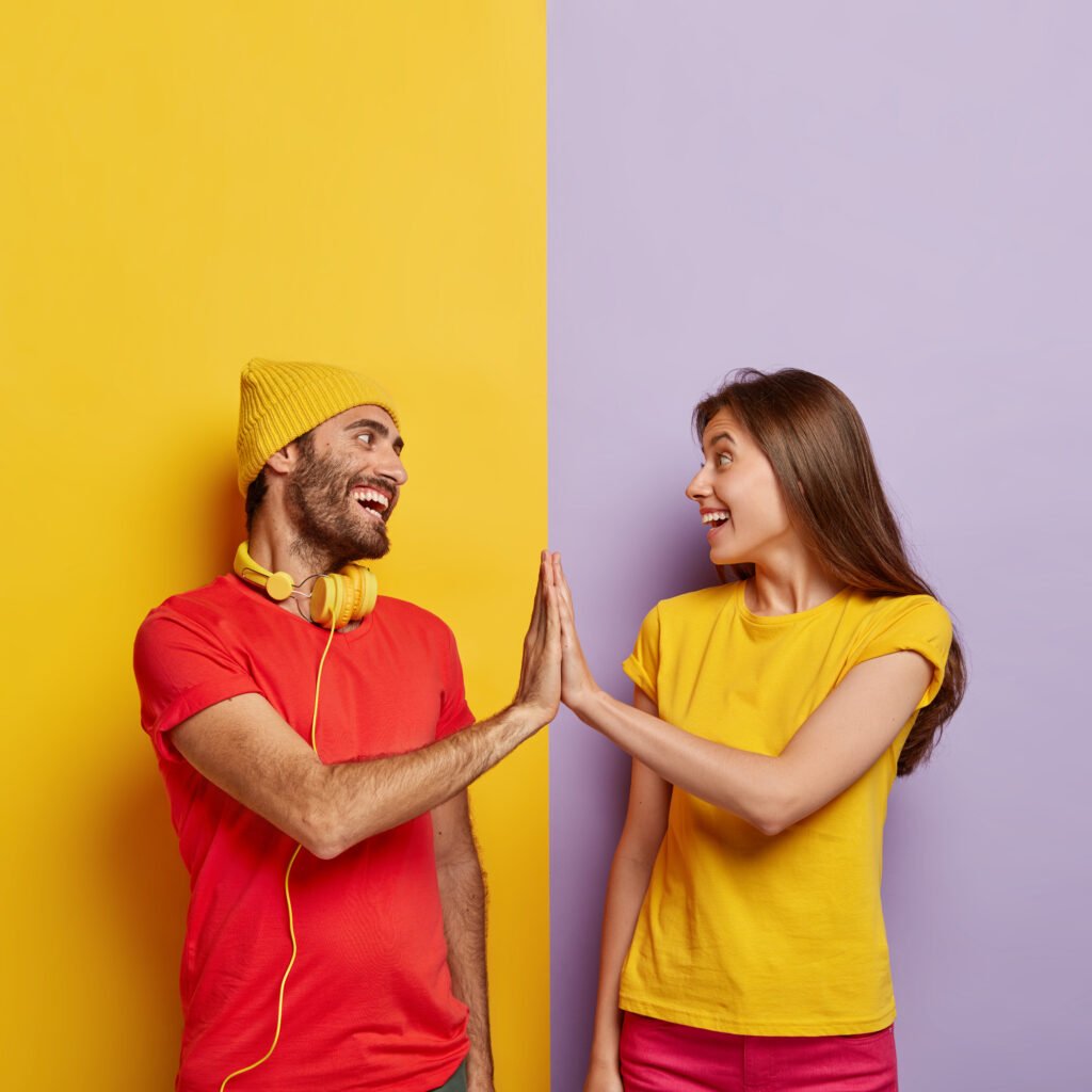 glad boyfriend and girlfriend touch by their palms, smile positively, agree about something, dressed in red and yellow casual t shirts, enjoy free time together, stand against colored background